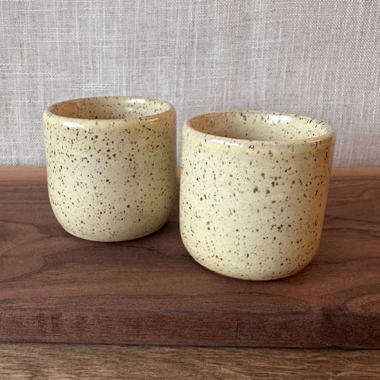 Two ceramic cups with speckled glaze on a wooden surface with a textured gray background