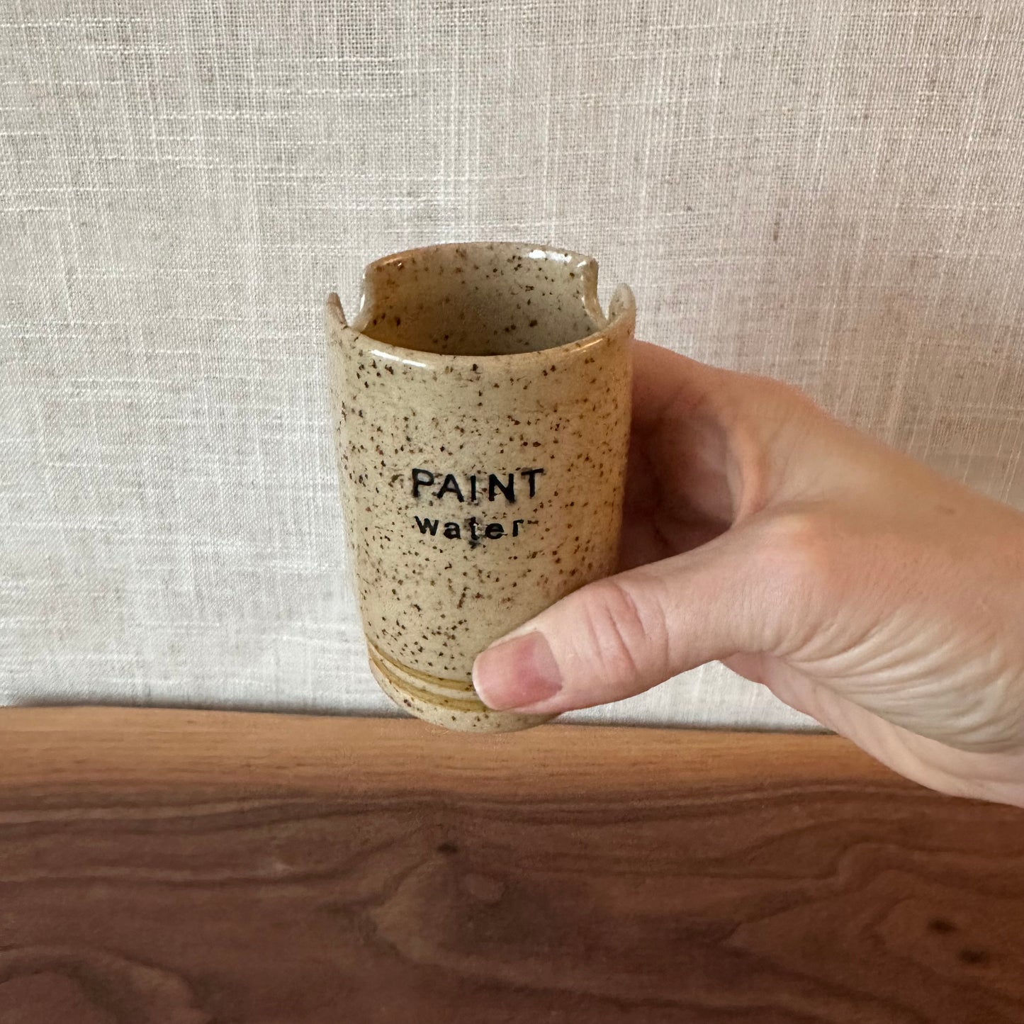 Hand holding a speckled ceramic cup with 'PAINT water' text against a textured beige wall.