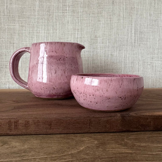 Pink ceramic pitcher and bowl on a wooden surface with a textured gray wall background