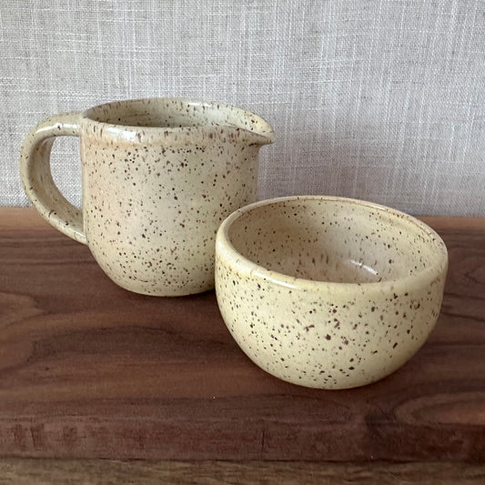 Speckled ceramic pitcher and bowl on a wooden surface with a textured gray background