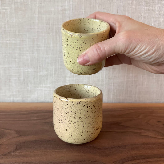 Hand holding a beige ceramic cup with speckled texture on a wooden surface.