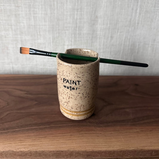 Speckled ceramic cup with 'PAINT water' text and a paintbrush on a wooden surface.