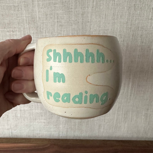 Mug with 'Shhhhh... I'm reading' text held by a hand against a neutral background