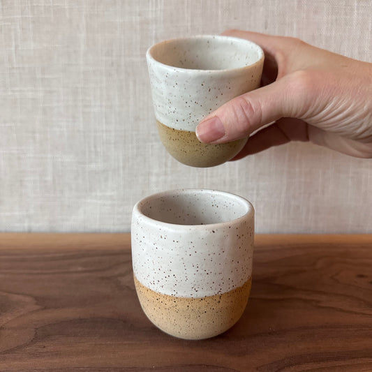 Hand holding a ceramic cup with another cup on a wooden surface against a neutral background