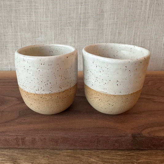 Two ceramic cups with speckled glaze on a wooden surface with a textured gray background